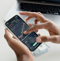 Hände halten ein Smartphone mit einer Finanz-App, die Börsenkurse zeigt. Im Hintergrund ein Laptop und Geldscheine.