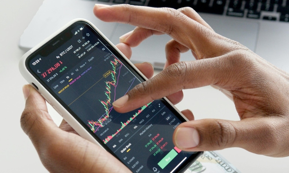 Hände halten ein Smartphone mit einer Finanz-App, die Börsenkurse zeigt. Im Hintergrund ein Laptop und Geldscheine.
